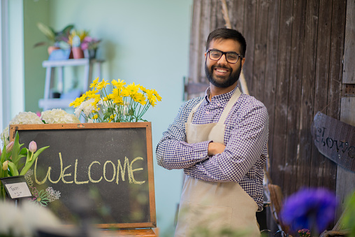 A male small business owner and entrepreneur is standing at the front of his florist shop by a welcome sign and is smiling while looking at the camera.
