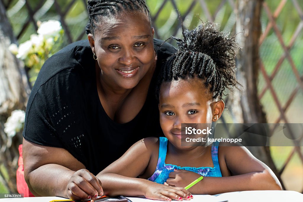 Portrait of african mother and little daughter in garden. Close up portrait of african mother and little daughter with braided hairstyle in garden.Girl drawing on paper with color pencils outdoors. Teacher Stock Photo