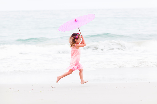 Happy young girl, holding a pink umbrella, running on the beach.