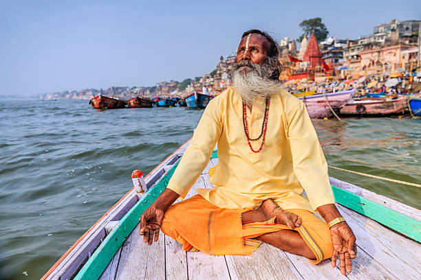 Sadhu is meditating in boat on Holy Ganges River, Varanasi In Hinduism, sadhu, or shadhu is a common term for a mystic, an ascetic, practitioner of yoga (yogi) and/or wandering monks. The sadhu is solely dedicated to achieving the fourth and final Hindu goal of life, moksha (liberation), through meditation and contemplation of Brahman. Sadhus often wear ochre-colored clothing, symbolizing renunciation. ghat photos stock pictures, royalty-free photos & images