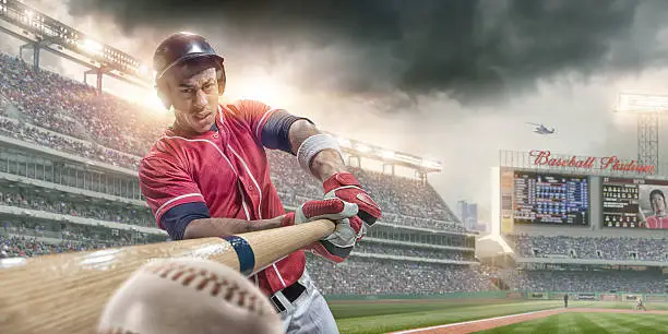 An extreme close up image of professional male baseball player in red uniform, just about to make contact and hit baseball. The action takes place during a baseball game in a generic outdoor floodlit stadium full of spectators under a dramatic stormy evening sky. The location is fake. 