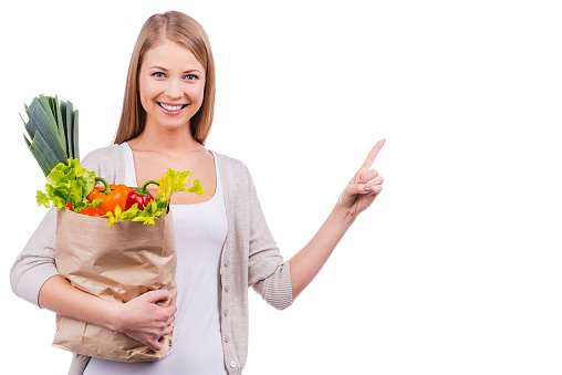 Beautiful young woman holding a shopping bag full of groceries and pointing away while standing against white background