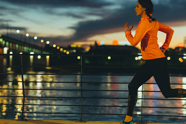 Young Woman Jogging Near River at night. She is wearing modern sporty clothing. With bridge and cityscape in background.