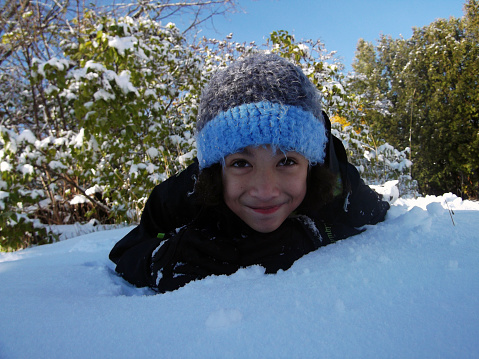 A cute tan Asian girl in a coat and hat, smiling and laying on her stomach in a pile of fresh snow.