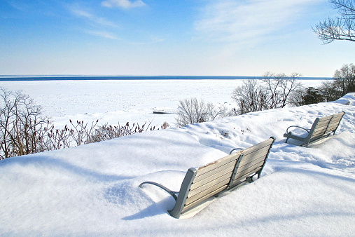 An afternoon view of snow covered park benches overlooking a snow and ice covered lake.
