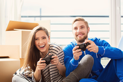 Portrat of happy young people during moving house, sitting on the floor in apartment and playing video games using gamepads. 
