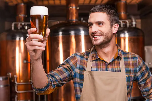 Happy young male brewer in apron holding glass with beer and looking at it with smile while standing in front of metal containers