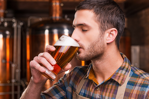 Handsome young male brewer in apron tasting fresh beer and keeping eyes closed while standing in front of metal containers
