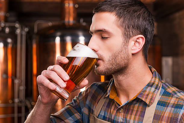 Tasting fresh brewed beer. Handsome young male brewer in apron tasting fresh beer and keeping eyes closed while standing in front of metal containers microbrewery photos stock pictures, royalty-free photos & images