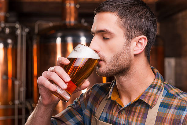 Tasting fresh brewed beer. Handsome young male brewer in apron tasting fresh beer and keeping eyes closed while standing in front of metal containers drinking beer stock pictures, royalty-free photos & images