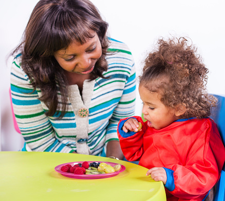 A portrait of an African American carer supervising a toddler during her meal.