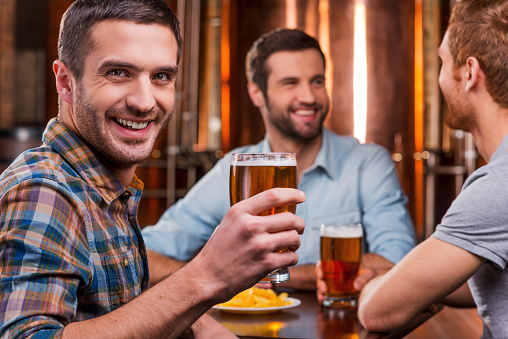 Handsome young man toasting with beer and smiling while sitting with his friends in beer pub