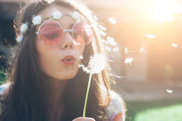 Young hippie woman blowing dandelion outside Close up of young hippie woman with pink sunglasses, wreath and long hair blowing dandelion in back yard. Sunlight and house view on background. hippie photos stock pictures, royalty-free photos & images