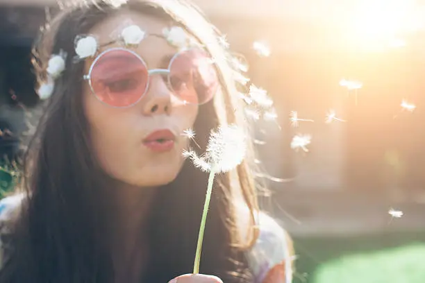 Close up of young hippie woman with pink sunglasses, wreath and long hair blowing dandelion in back yard. Sunlight and house view on background.
