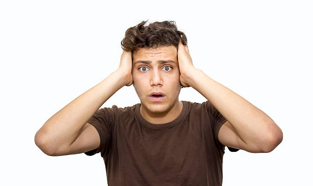 Portrait of shocked young man over white background Portrait of shocked young man over white background. Frustrated teenage boy looking at camera with negative facial expression. Worried boy has got short, curly brown hair. Horizontal composition. Studio shot. Worried young man's hands on his head. confusion raised eyebrows human face men stock pictures, royalty-free photos & images