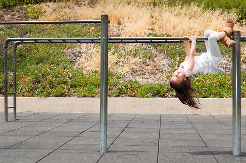 A pretty daring and adventerous 6 years old girl is hanging upside down on monkey bars in a playground. In the background, rubber floor tiles can be seen, which ensure playground safety, to prevent injury from falls.