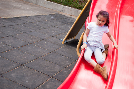 A pretty 6 years old girl is sliding down a red slide in a playground. In the background, rubber floor tiles can be seen, which ensure playground safety, to prevent injury from falls.