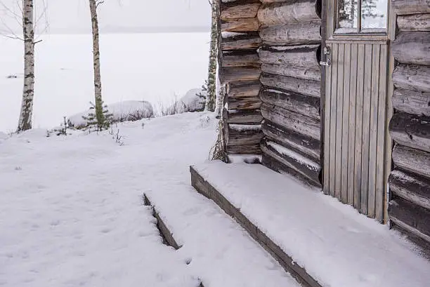 Small and old log cottage in winter. Frozen lake behind the cottage.