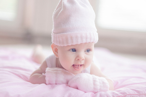 Adorable playfull baby girl, lying at her tommy on a pink comforter on the floor. She's playing wit her mittens laughing,