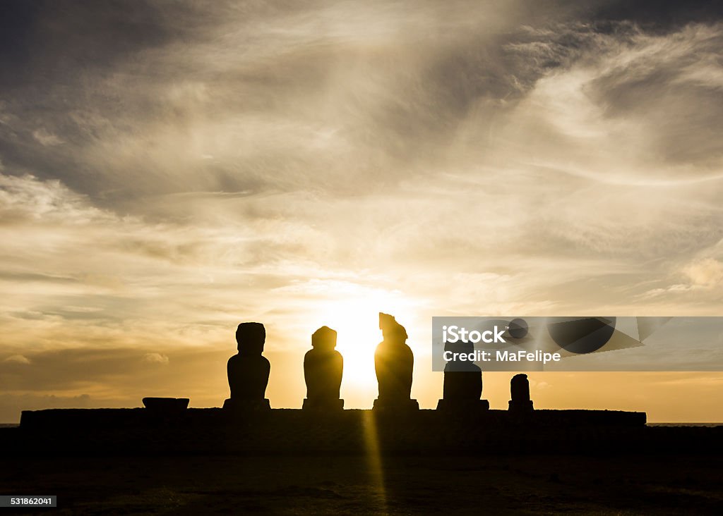 Ahu Tahai Moai Statues at Sunset, Easter Island A DSLR photo of the five Moai statues in Ahu Tahai, Easter Island. They are silhouetted against the bright yellow setting sun exactly behind them. The sky is filled woth many sparse white clouds.There is no one around. 2015 Stock Photo