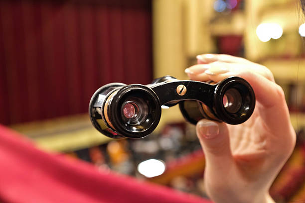 A set of theater binoculars in a female hand. The theater binoculars are black, grey and golden. The two eye pieces are on the ends of short tubes. The two tubes are connected in the center with a hinge. An adjustment wheel is in the center. The background is out of focus and depicts the inside of a theater. The corner of the stage is visible.