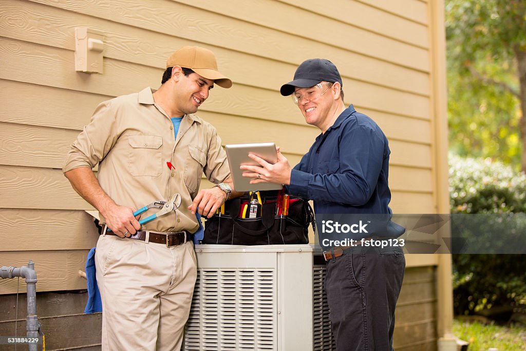 Air conditioner repairmen work on home unit. Blue collar workers. Caucasian and Latin descent repairmen work on a home's air conditioner unit outdoors. Manager explains job details using a digital tablet. Other man is checking unit using the tools in his tool bag.  They both wear uniforms.  Repairmen, electricians, inspectors, service industry workers.  Air Conditioner Stock Photo