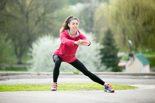 Portrait of sporty woman doing stretching exercises in park before training. Female athlete preparing for jogging outdoors. Runner doing side lunges. Sport active lifestyle concept. Full length