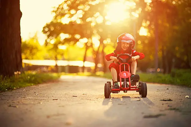 Happy little boy aged 6 is driving a go-kart (or kart, soapbox car, cyclekart). Road and sunset visible in the background. The boy is wearing a helmet and racer outfit.