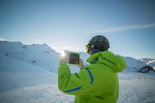 Young man at the top of the ski runs taking a picture of the snow capped alps with a digital tablet.Switzerland.