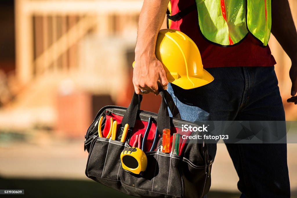 Construction worker working at a job site. Toolbag, hardhat. A construction worker busy working at a job site. He holds a tool box full of tools and a hard hat. Framed house, building in background. He is wearing a safety vest. Work Tool Stock Photo Construction worker working at a job site. Toolbag, hardhat. A construction worker busy working at a job site. He holds a tool box full of tools and a hard hat. Framed house, building in background. He is wearing a safety vest. Work Tool Stock Photo