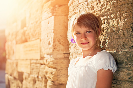 Portait of a little girl next to a thousand years old wall. Little girl is aged 7 and is wearing a white dress. The girl is visiting town of Alcudia, Mallorca.