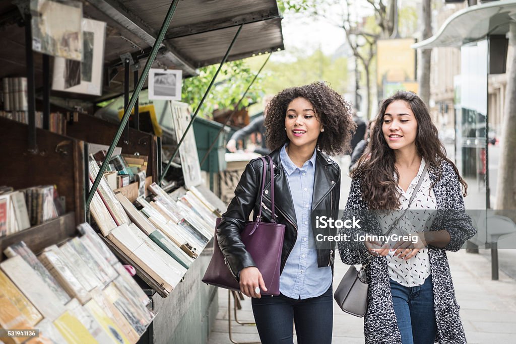 Two female friends walking past bookshop Two young women looking at second hand books in an outdoor book store. Two female friends out shopping in Paris, France Paris - France Stock Photo
