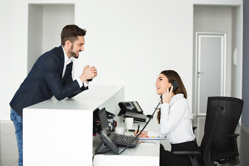 Young smiling female secretary is talking on the phone while communicating with cheerful businessman who is leaning against the receptionist desk.
