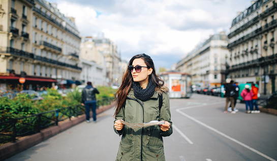 Vintage toned image of a young woman tourist in Paris, holding a map, looking around. She is wearing a fashionable olive green parka jacket and a scarf, standing on the streets in the Paris city center. Shot with wide angle large aperture prime lens for more shallow depth of field.