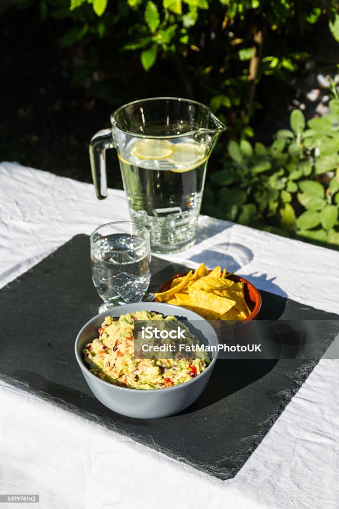 Guacamole in bowl with tortilla chips and iced water. Outdoors Home made guacamole in ceramic bowl and tortilla chips and soured cream on the side with jug and glass of iced water. On slate mat, outside on linen covered table. Evening light. Avocado Stock Photo