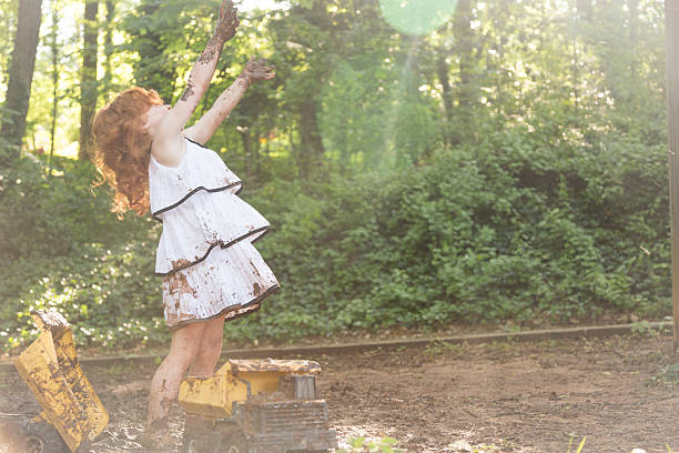 A little girl plays in the mud.  She is wearing a white dress with black stripes.  She has red hair and caucasian skin.   She is splashing and playing in the mud with toy trucks and is very messy and dirty.  She is having a good time and is expressing positivity and happiness.