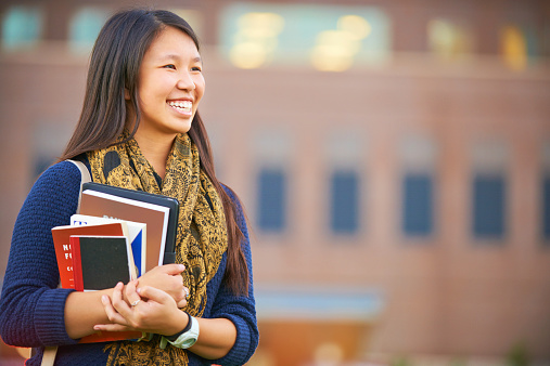 Portrait of happy student girl with cellphone and workbooks walking in college campus and smiling at camera, having break after classes outdoors, copy space