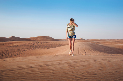 Tanned young girl in denim shorts and a patterned blouse is walking alone on the sand dunes of the desert