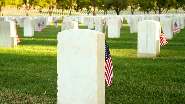 Dolly camera moves up past a marble tombstone at a US National Cemetery. Many more tombstones can be seen in the background. Small flags are placed next to each tombstone.