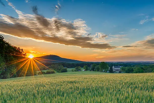 A perfect Spring day ends as the sun sets and sprays warm golden light over a beautiful farm with fields sprouting, a stone house, and mountain ridges in the background.