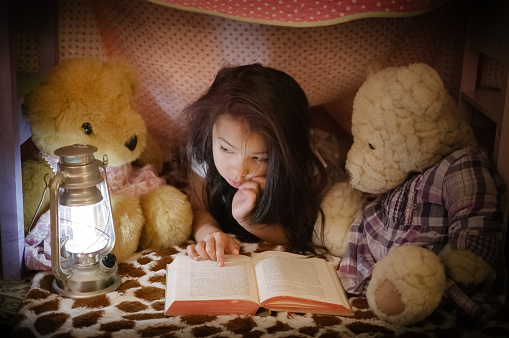 A pretty little girl is reading a book to her teddy bears. They are all hiding inside a fort made of chairs and blankets. An electric lantern is lighting up the interior of the fort, and the pages of the book she's reading.
