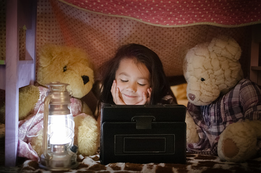 A pretty little girl is using an tablet computer / tablet. They are all hiding inside a fort made of chairs and blankets. An electric lantern is lighting up the interior of the fort.