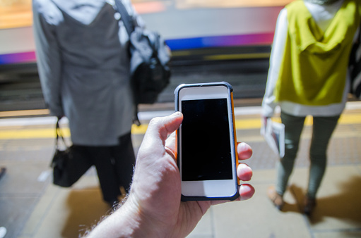 Close up on a cell phone held in left hand in metro or subway  with people waiting on dock in background  (POV)