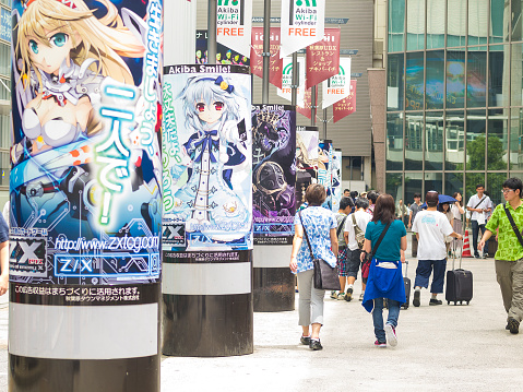 Tokyo, Japan - August 17, 2014: Back view of people walking on pavement near Akihabara train station. On left side are columns with large paper posters with the characters from the Japanese comic book heroes and computer animations. In background is big, modern building.