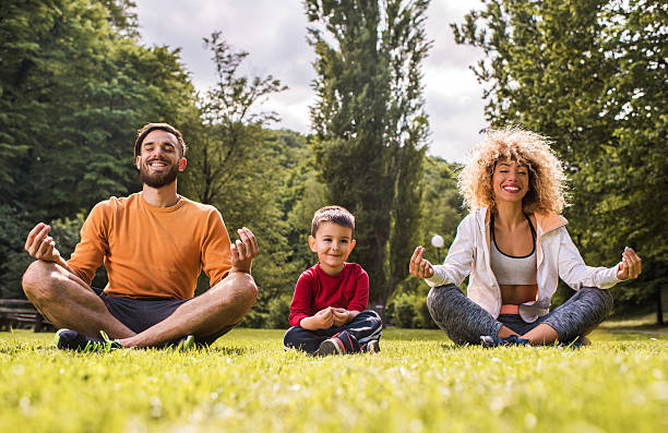 Jovem família feliz praticando exercícios de relaxamento de ioga na grama. Pais felizes praticando ioga na posição de lótus enquanto o filho está sentado entre eles, olhando para o outro lado. O foco está no menino. Fotos de homem meditando, fotos e imagens livres de direitos autorais