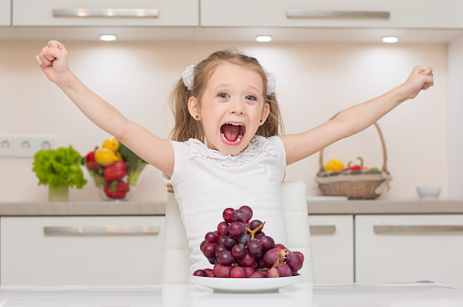 Sweet cute young positive girl with two ponytails shouts and put hands up near bowl full of fresh grapes.Little cute preschool girl in the kitchen smiles and hugs bowl full of healthy fresh vegetables - broccoli, paprika, lemons