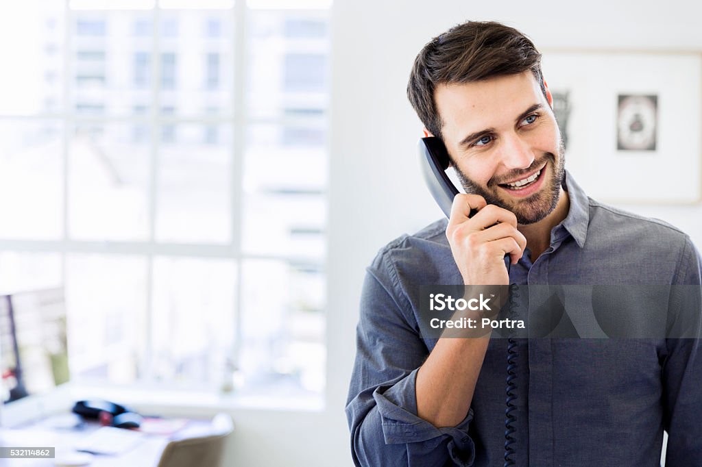 Happy businessman using landline phone in office A photo of happy businessman using landline phone in office. Smiling male professional is looking away while talking through telephone receiver. Handsome executive is at brightly lit workplace. Landline Phone Stock Photo