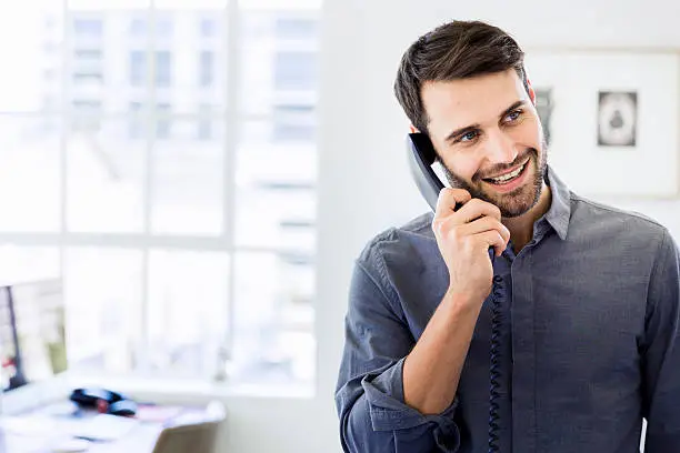 A photo of happy businessman using landline phone in office. Smiling male professional is looking away while talking through telephone receiver. Handsome executive is at brightly lit workplace.