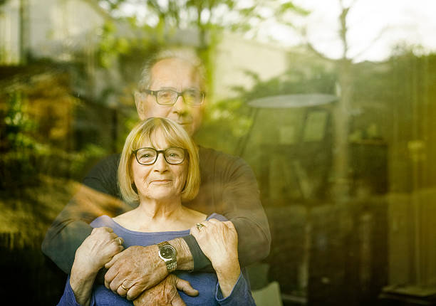Loving senior couple seen through glass window A photo of loving senior couple seen through glass window. Elderly male is embracing female partner from behind. They are spending quality time together at home. 60 69 years stock pictures, royalty-free photos & images