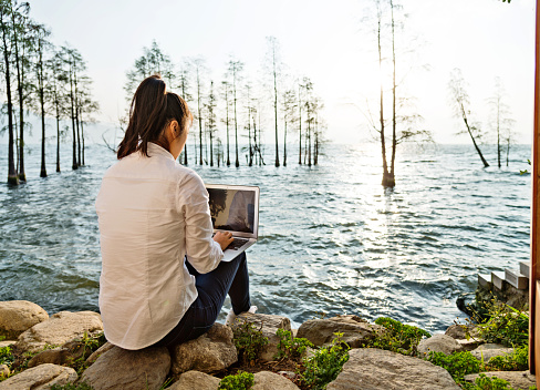 Young woman working on her laptop while sitting by the lake.